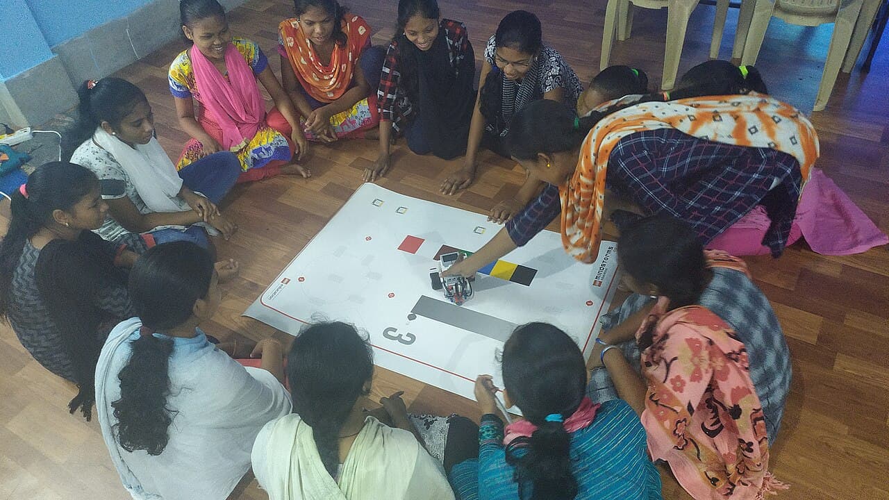 Students gathered in a circle around a mat, programming and testing a small robot together